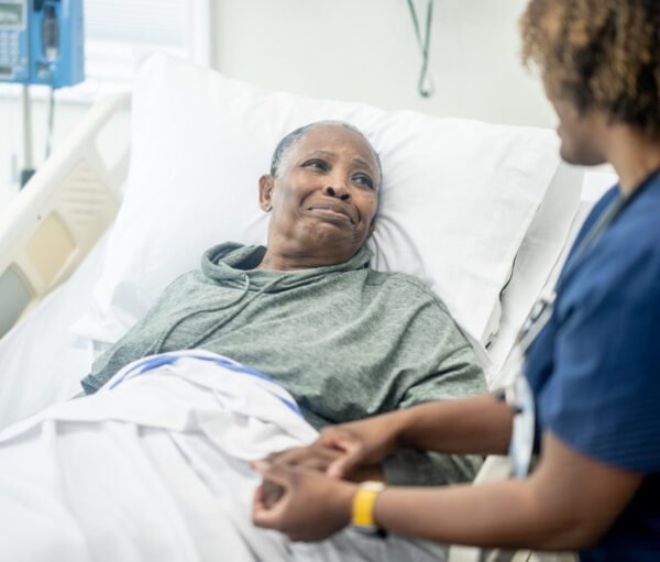 A female nurse of African decent checks in on her senior patient as she awaits to be called for surgery. She is smiling as she reassures her patient and puts her at ease before the procedure.