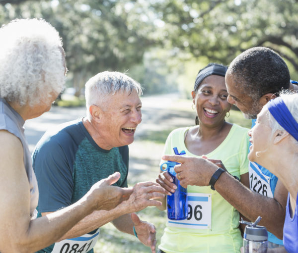 A group of five multi-ethnic senior men and women wearing marathon bibs, hanging around, talking and smiling, holding water bottles, at the end of their race.